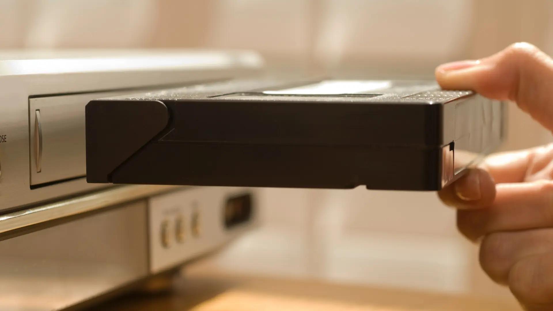 A hand sliding a black VHS cassette into the front slot of a silver video recorder