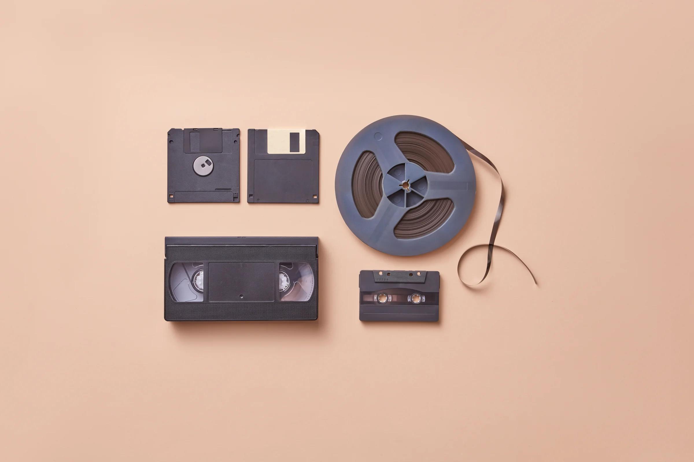 An open reel of brown audio tape with the tape end trailing, beside a VHS cassette, an audio cassette and two floppy disks on a peach background